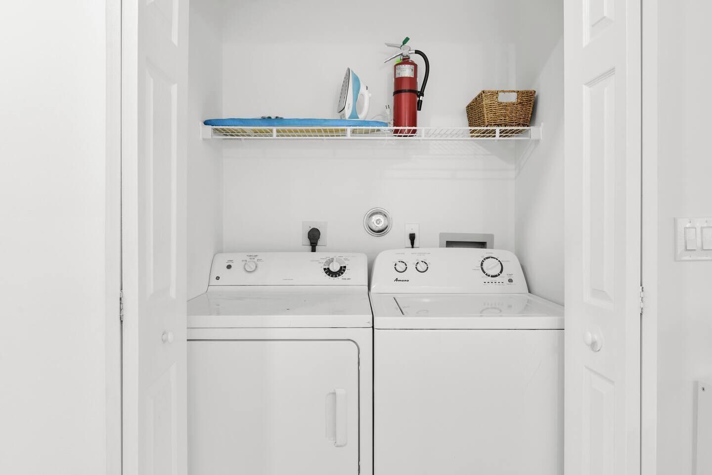 A laundry room with two white front-loading washing machines, a red fire extinguisher, and a wicker basket on a shelf.