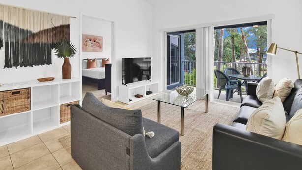 A modern living room with a sofa, TV, and glass coffee table. There is a balcony with outdoor furniture and a view of trees. - 350D - Boca Retreat w/ Pool, Parking, Near Evert Tennis & Shops (Boca Raton)