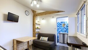 Et kompakt værelse med sofa, lille bord, fastgjort fjernsyn, vækkeur og flisebeklædt bagpanel.