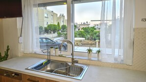 Une cuisine dotée d’un évier en acier inoxydable, d’une fenêtre ornée de rideaux blancs et offrant une vue sur un quartier résidentiel avec des maisons et une voiture.
