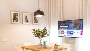 A modern dining area with a wooden table and white chairs, a hanging pendant light, framed pictures on the wall, and a TV displaying a Netflix welcome screen.