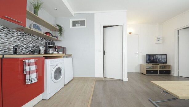 A modern kitchen with a washing machine, a coffee maker, and a microwave. There is a TV in the living area and a door leading to another room.