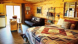 A cozy log cabin bedroom with a large bed, a wooden dresser, a small table with stools, and a window with curtains.