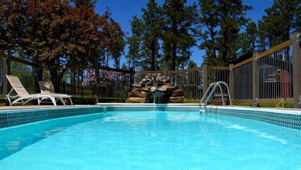 A clear blue pool with a rock waterfall feature, surrounded by a wooden fence and trees.