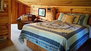 Un acogedor dormitorio de cabaña de madera con techo de madera, una cama con colcha estampada, una mesita de noche de madera y una silla con cojines.