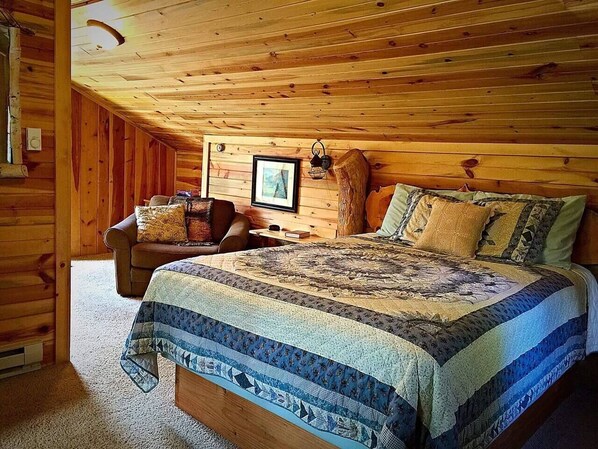 Un acogedor dormitorio de cabaña de madera con techo de madera, una cama con colcha estampada, una mesita de noche de madera y una silla con cojines.