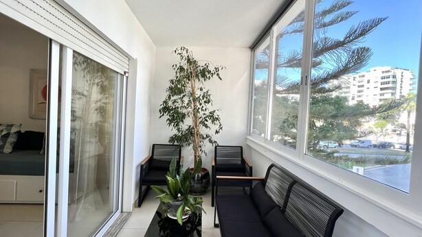 Ein sonnendurchflutetes Zimmer mit einer schwarzen Couch, einem Glastisch mit Pflanzen und Blick auf eine Stadtlandschaft durch große Fenster.
