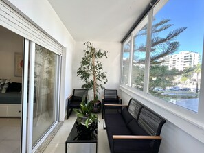 A sunlit room with a black sofa, a glass table with plants, and a view of a cityscape through large windows.