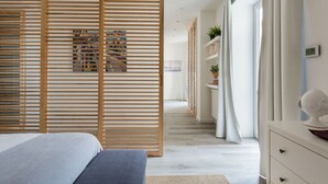 Ein modernes Schlafzimmer mit einer Holztrennwand, einem Bett mit grau-blauer Bettdecke, einer weißen Kommode und einem Blumentopf auf der Kommode.