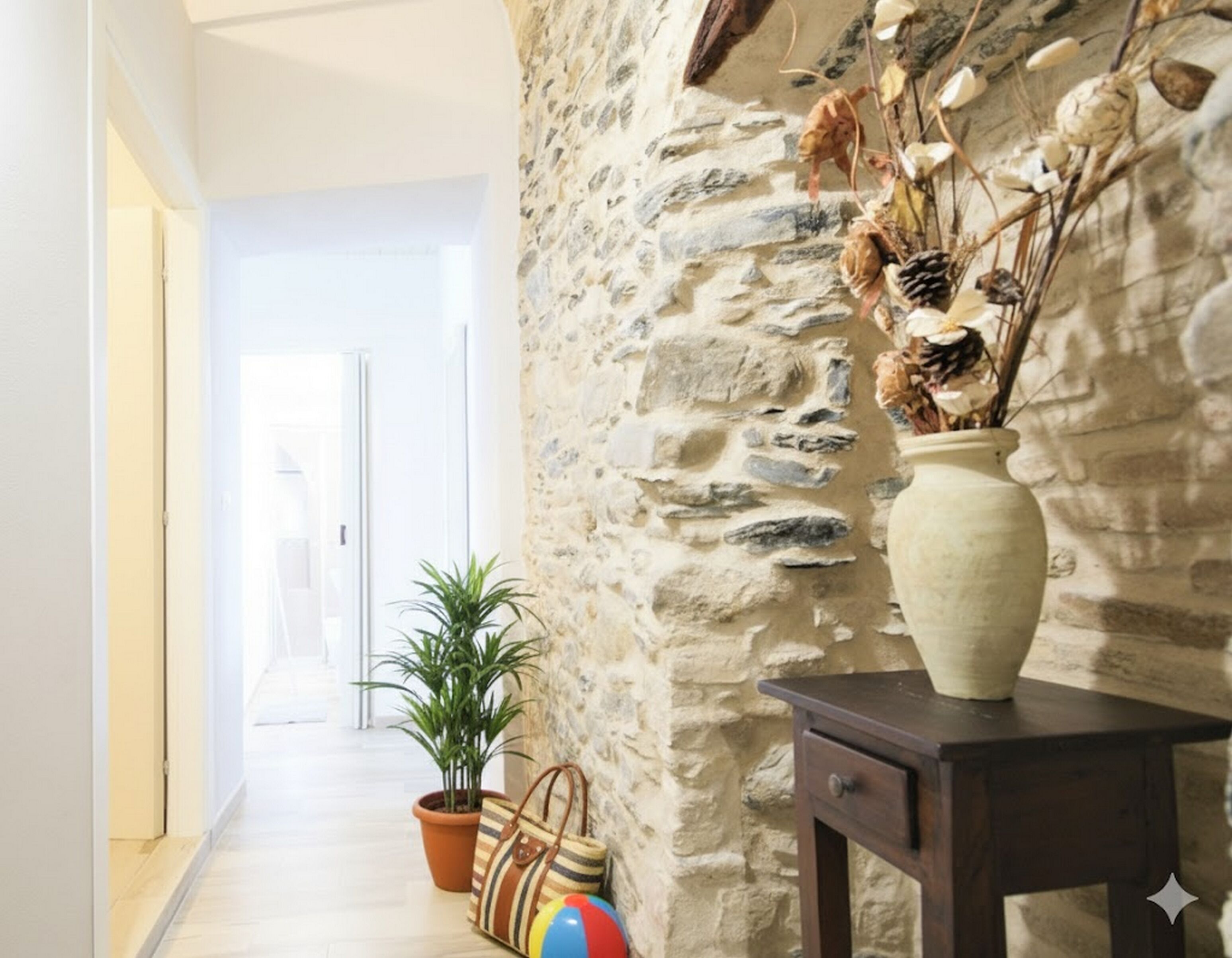 Un couloir avec un mur en pierre, une plante en pot, un sac à rayures, un ballon de plage et un vase contenant des branches séchées.
