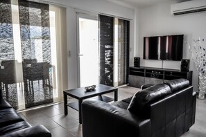 Relax in this stylish living room with a black leather sofa, flat-screen TV, and sliding glass door leading to a balcony with a tree view.