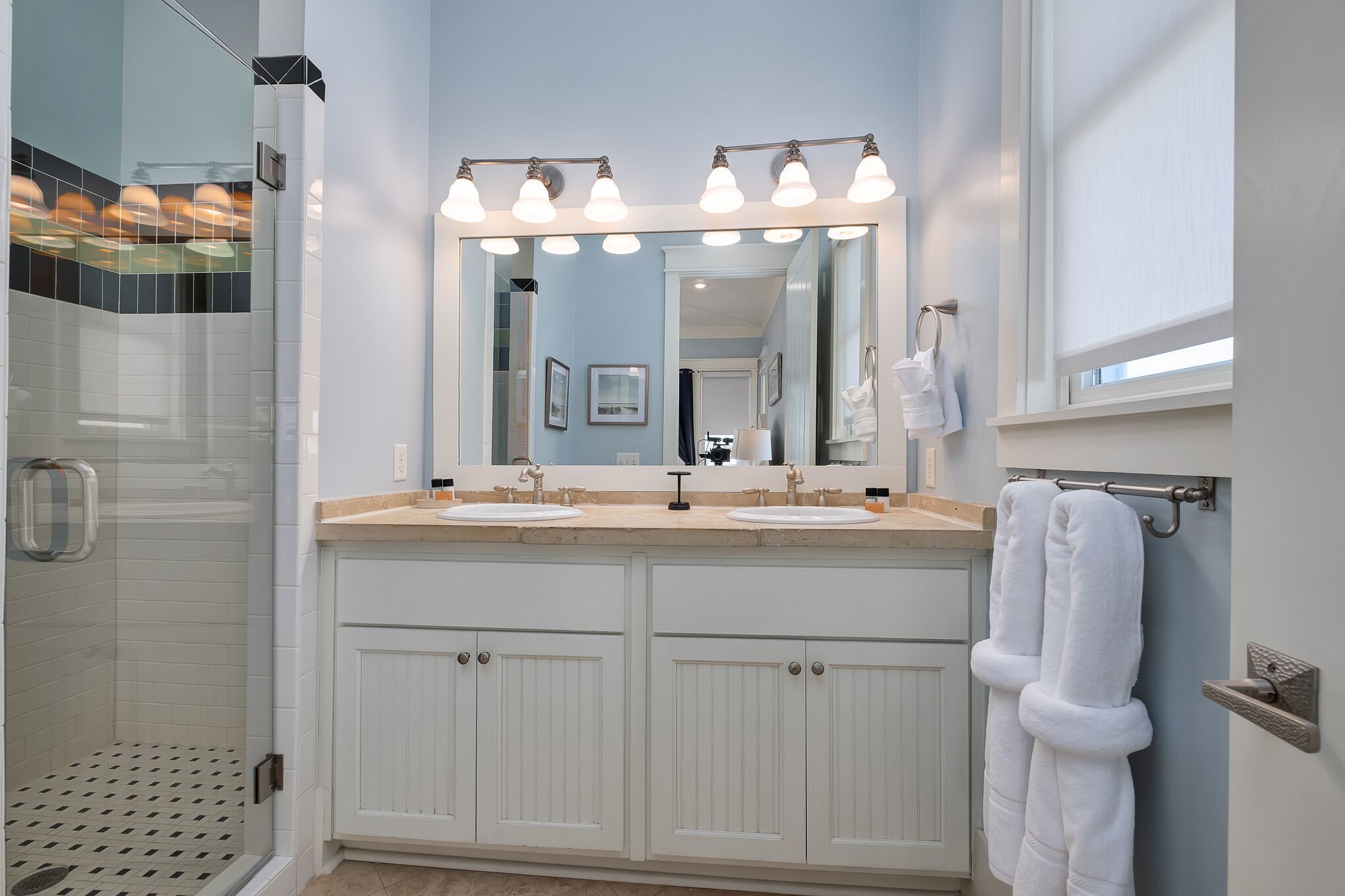 A bathroom with a double sink vanity, a large mirror with lights, a shower area, and a towel rack.