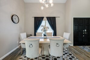 A dining room with a wooden table, four upholstered chairs, a wall clock, a chandelier, and a window with dark curtains.