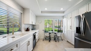 A modern kitchen with white cabinetry, stainless steel appliances, a dining table with chairs, and a view of a green landscape through the window. - Santa Rosa Cove #6 , 3 bedroom 2 bath Condo! (La Quinta)