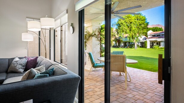 A modern living room with a grey sofa, a glass door leading to a patio with a table and chairs, and a view of a well-maintained garden and buildings in the background. - Santa Rosa Cove #6 , 3 bedroom 2 bath Condo! (La Quinta)