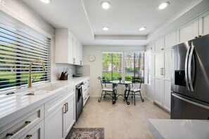 A modern kitchen with white cabinetry, stainless steel appliances, a dining table with chairs, and a view of a green landscape through the window.