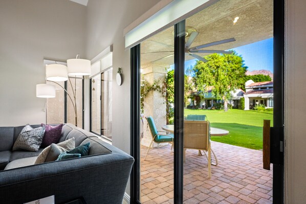 A modern living room with a grey sofa, a glass door leading to a patio with a table and chairs, and a view of a well-maintained garden and buildings in the background.