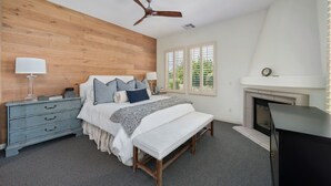 Une chambre à coucher avec une tête de lit en bois, un lit avec des oreillers, une table de chevet avec une lampe, un banc, une cheminée et un ventilateur de plafond.