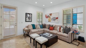 Un salon avec un canapé d’angle beige, une table basse et une table d’appoint avec une lampe. Il y a deux fenêtres équipées de stores, une porte avec un moustiquaire et un tableau abstrait encadré accroché au mur.