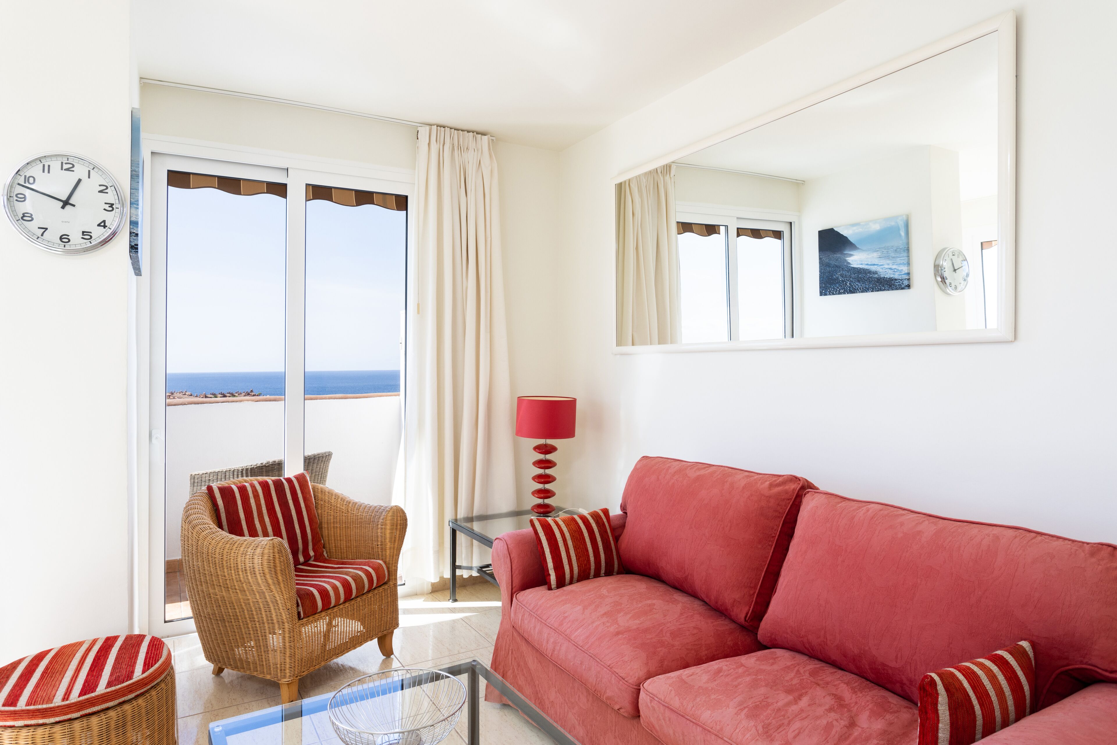 Uma sala de estar com um sofá vermelho, poltrona de vime e uma mesa de centro de vidro. Há uma janela grande com cortinas, um abajur vermelho e um relógio de parede.