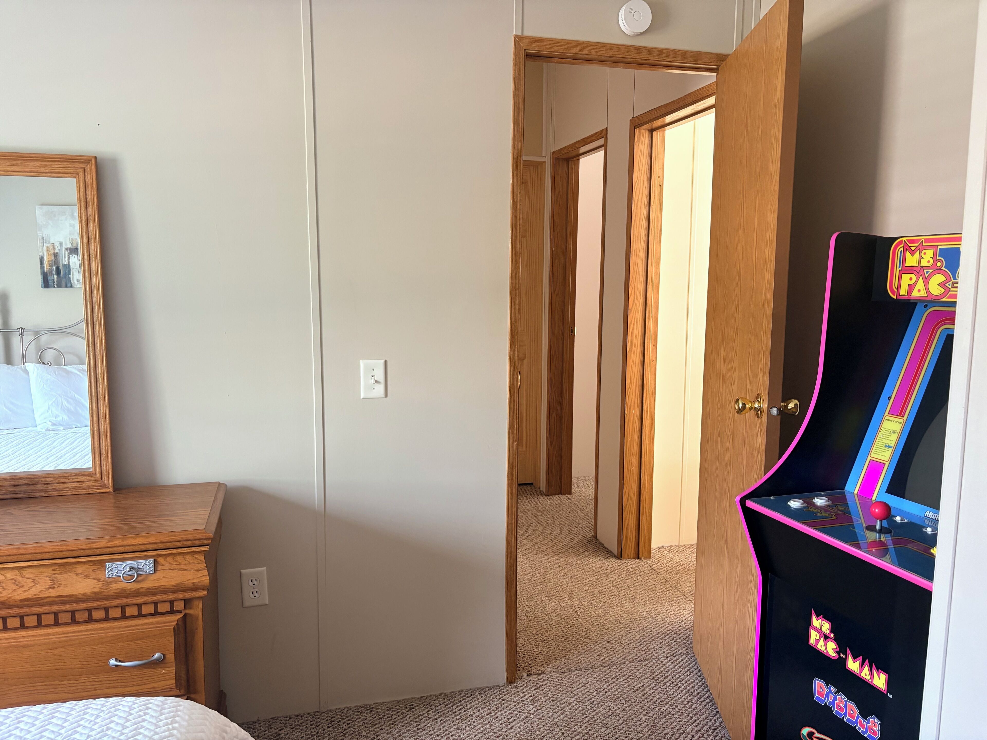 A bedroom with a wooden dresser, a mirror, a door leading to another room, and a Pac-Man arcade machine.