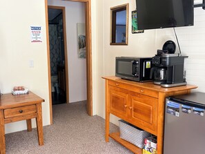 Eine Küchenzeile mit Mikrowelle, Kaffeemaschine und Kühlschrank. Ein Holzkörperschrank mit Schublade sowie ein kleiner Tisch mit einem gefalteten Tuch sind zu sehen. Ein Hinweisschild ist an der Wand angebracht.