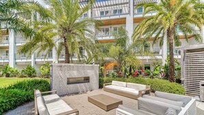 Un área de patio al aire libre con asientos y un hogar para fuego integrado, rodeada de abundante follaje y palmeras.