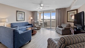 A hotel room with a blue sofa, a patterned armchair, a television, and a balcony with a view. - Yacht Club Villas #1-701 Great Location! (North Myrtle Beach)