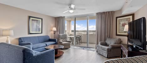 A hotel room with a blue sofa, a patterned armchair, a television, and a balcony with a view.