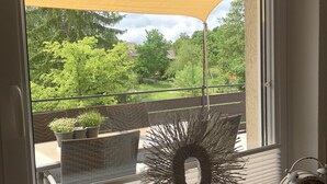A window with a view of a garden, a wicker chair, a decorative wreath, a clock, and a small white sculpture.