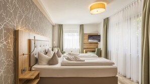 A modern hotel room with a large bed, wooden headboard, a bedside table, a TV, and a large window with curtains.