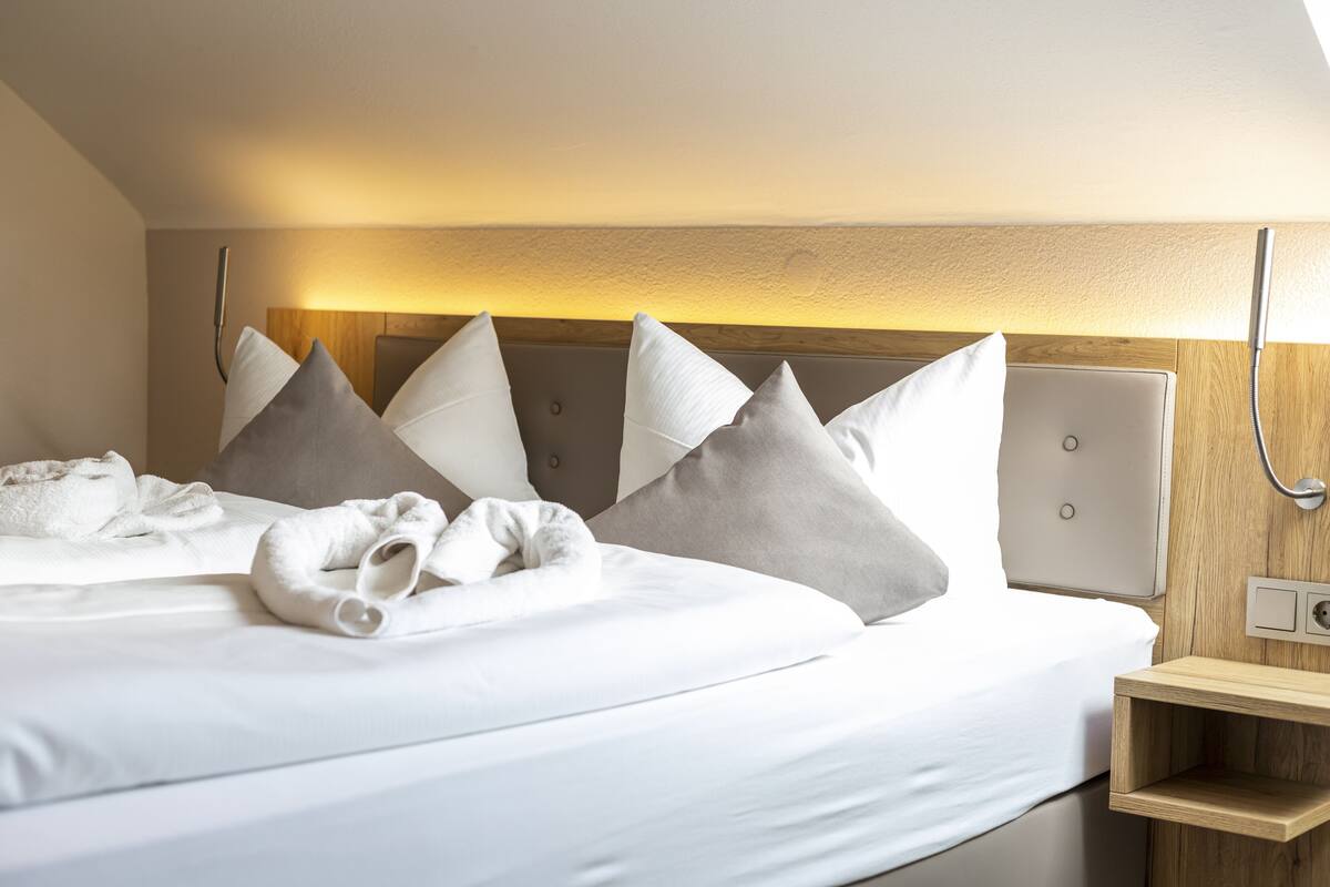 Una cama bien tendida con sábanas blancas y almohadas grises, un cabecero de madera y una mesita de noche con una lámpara.