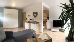 A modern living room with a sofa, coffee table, and a potted plant. There is a heart-shaped decoration on the wall and a television on a stand.