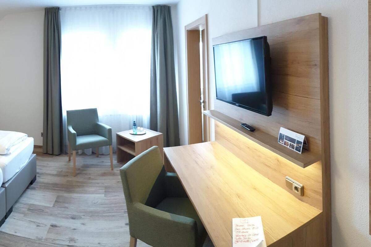Habitación de hotel con cama, escritorio, silla, televisor y ventana con cortinas.
