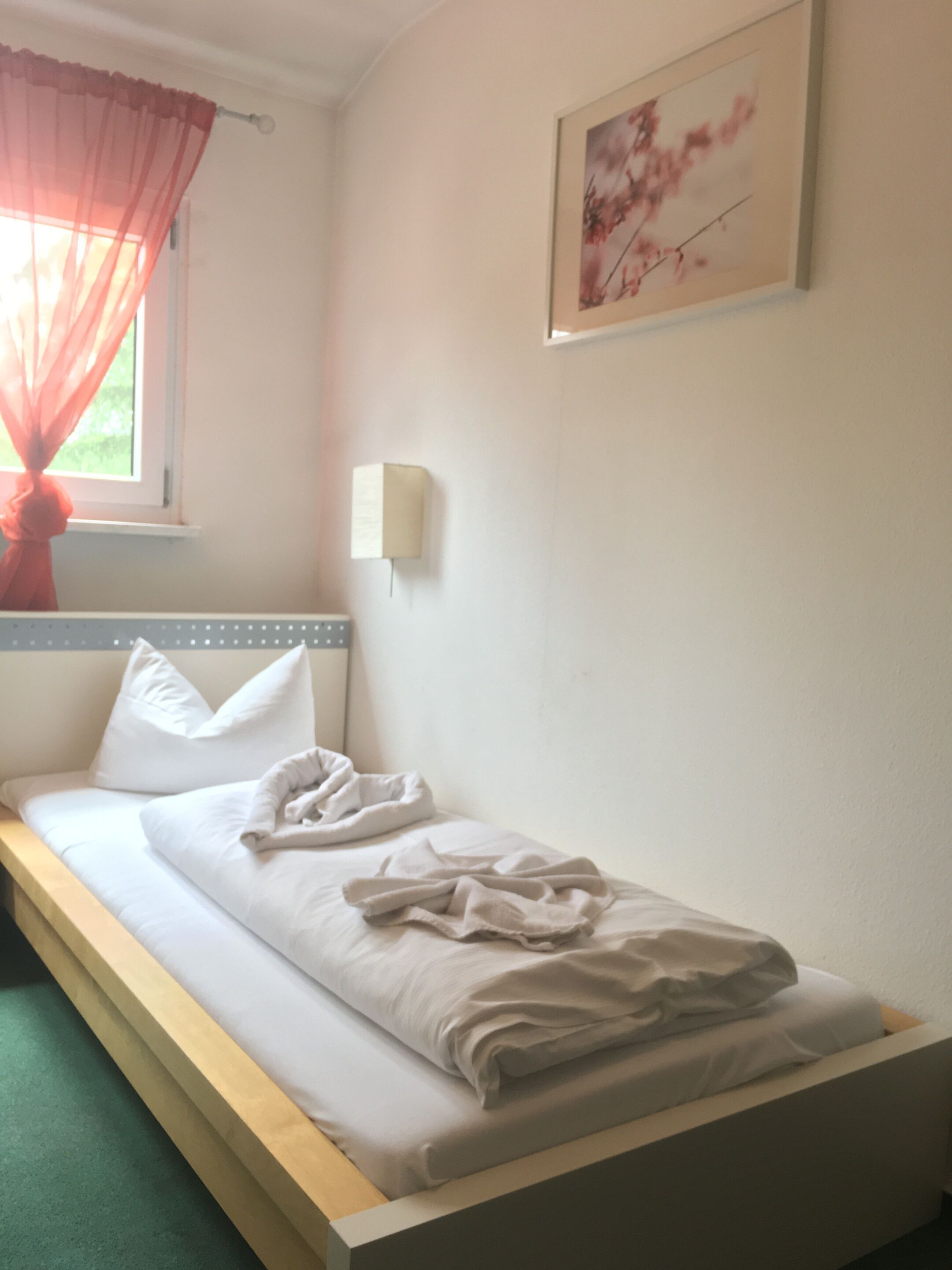 A single bed with white bedding, a window with red curtains, a wall-mounted light, and a framed picture on the wall.