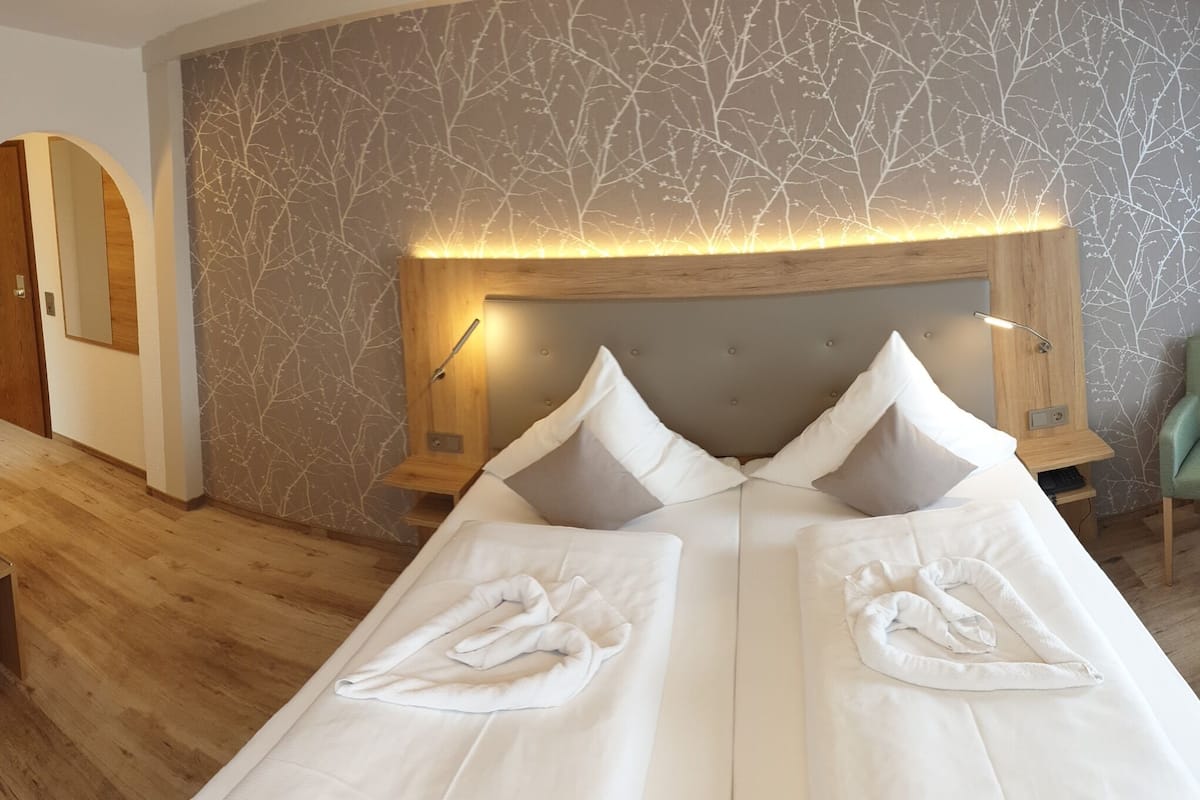 Habitación de hotel con una cama grande, dos almohadas, un cabecero de madera, una pared con diseño de árboles y una silla verde.