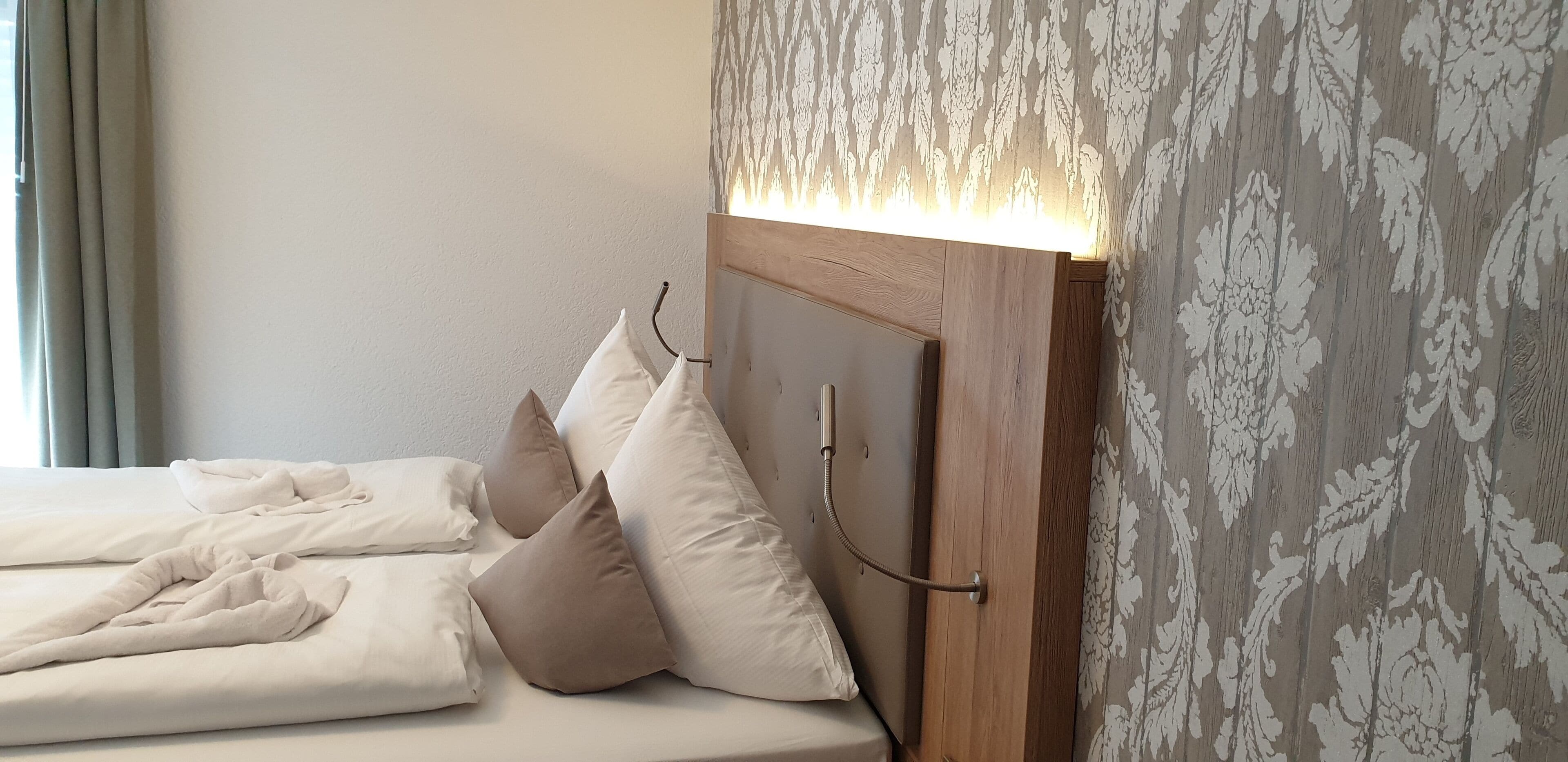 A neatly made bed with white and beige pillows, a wooden headboard, and a wall with a patterned wallpaper.