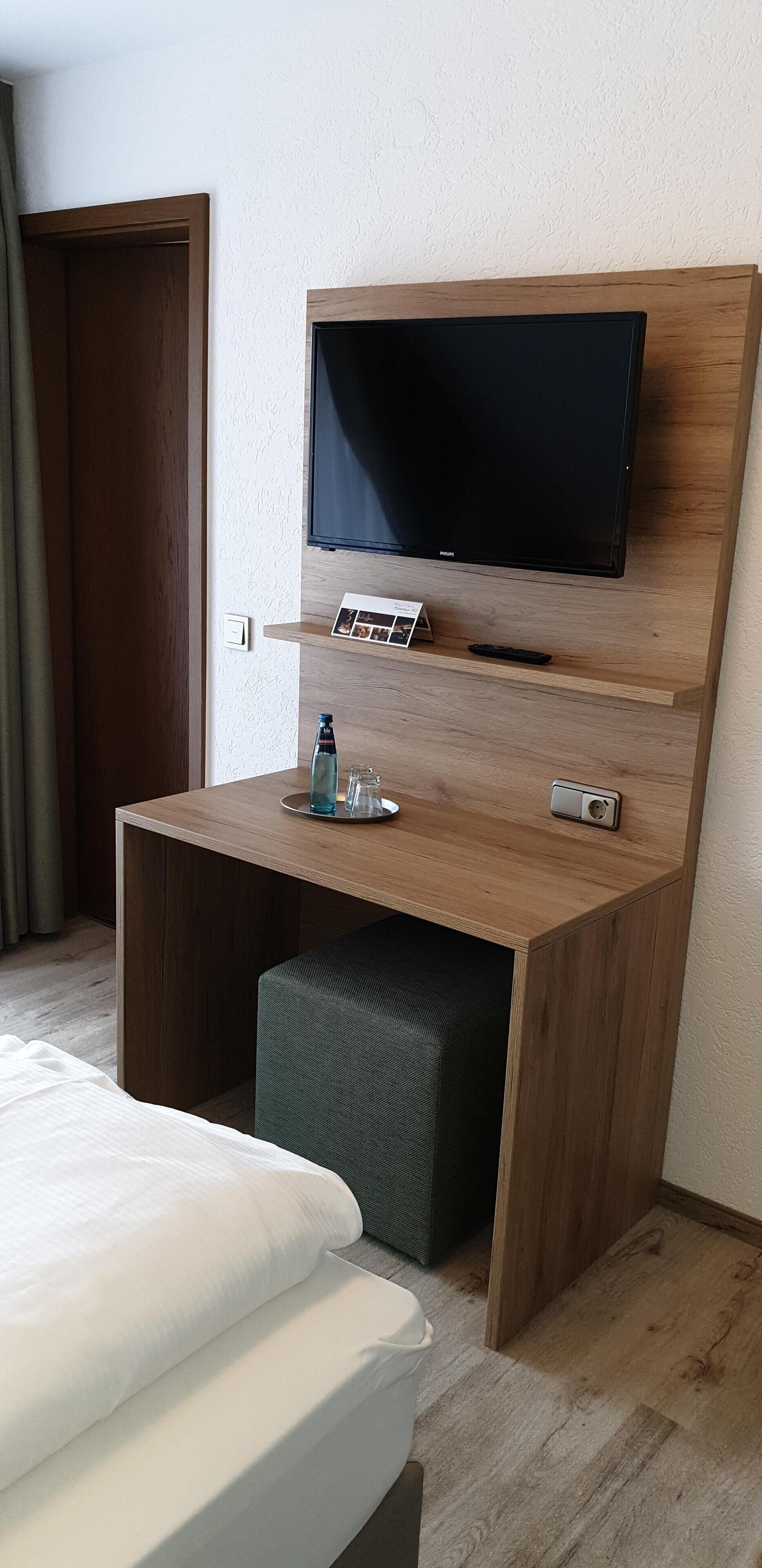 A hotel room with a wooden desk, a flat-screen TV, a small table with a remote control, a water bottle, and a glass.