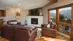 Ein Wohnzimmer mit Kamin, Ledersofas, einem Fernseher und einem großen Fenster mit Blick auf die Berge.