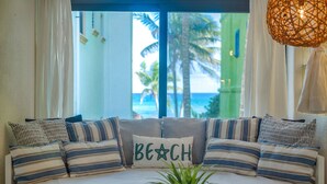 A cozy seating area with a sofa adorned with striped pillows and a 'Beach' pillow, positioned in front of a large window offering a view of palm trees and the ocean. - Affordable, bright studio with beach access, AC and WiFi! (Akumal)