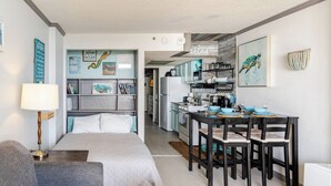 A compact, modern room with a bed, a desk with chairs, a kitchenette, and a bookshelf.