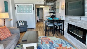 A modern living room with a sofa, coffee table, and a rug with a fish design. There is a kitchen area with a dining table and chairs, and a TV mounted on the wall.