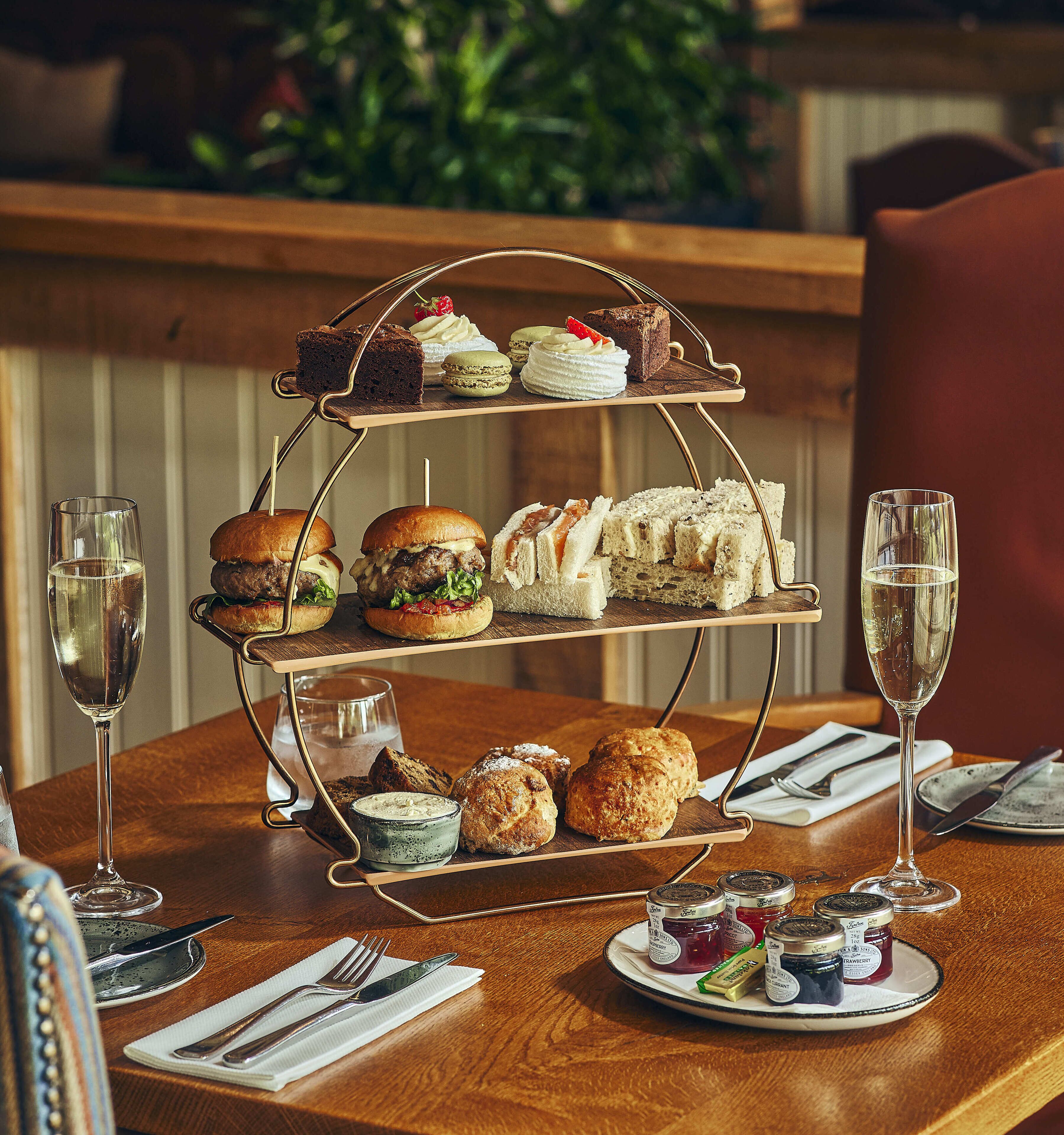 A three-tiered dessert stand with various pastries and sandwiches on a wooden table, accompanied by two glasses of champagne.