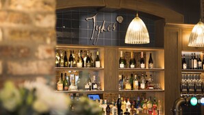 A bar area with shelves of liquor bottles and glasses, a sign above the shelves, and hanging pendant lights. - Sandburn Hall Hotel (York)