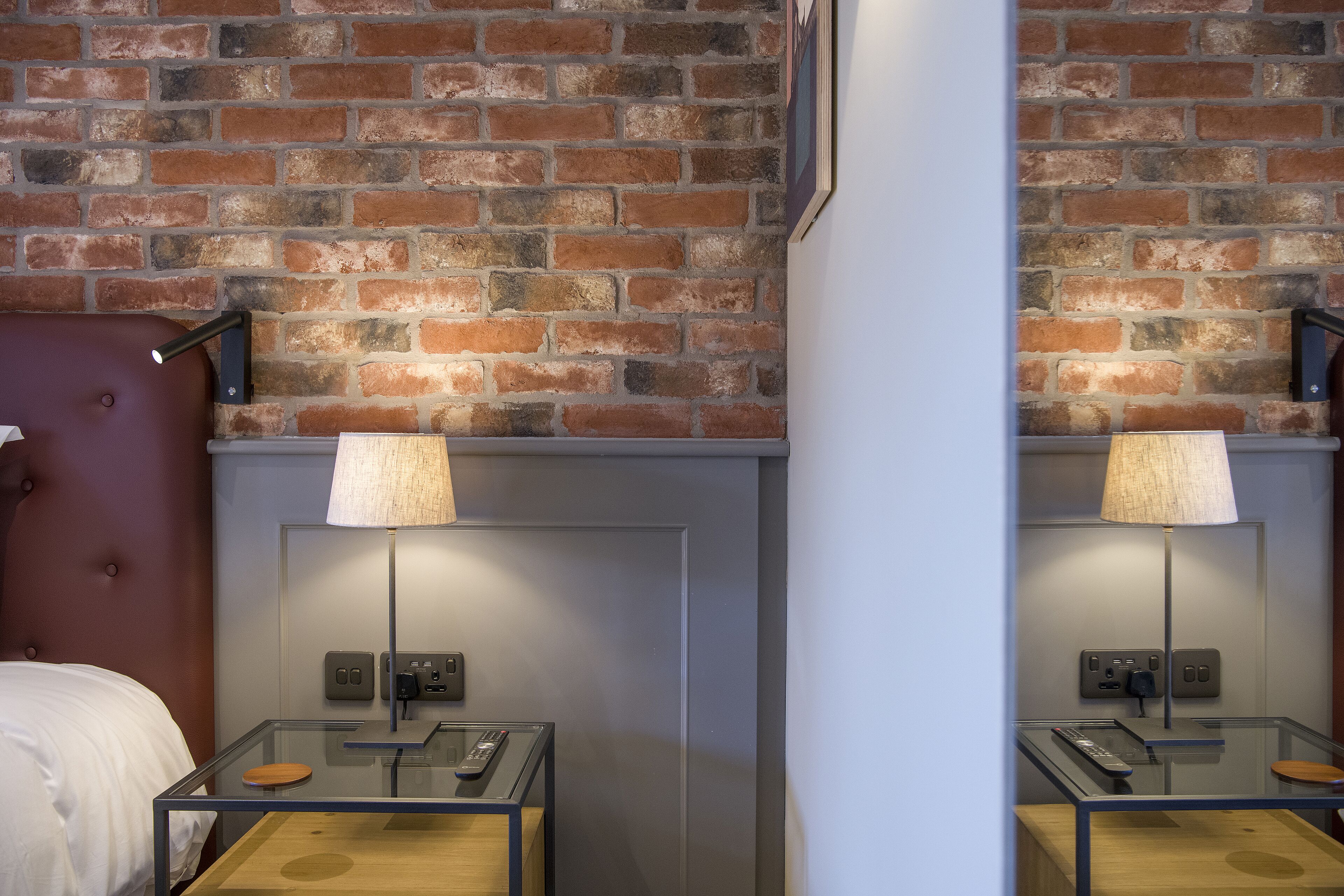 A hotel room with a brick wall, a bed with a red headboard, a glass table with a remote control, and a lamp.