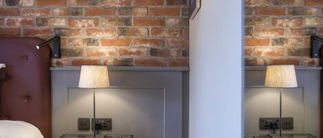 A hotel room with a brick wall, a bed with a red headboard, a glass table with a remote control, and a lamp.