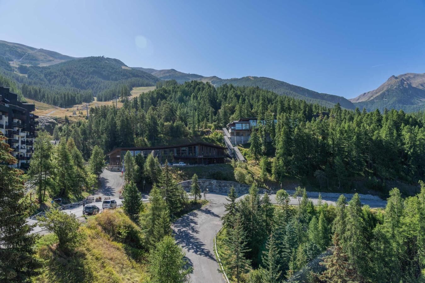 Un chalet avec une vue sur les montagnes, entouré d’une forêt dense, comprenant un espace de stationnement et un bâtiment principal doté d’un toit en pente.