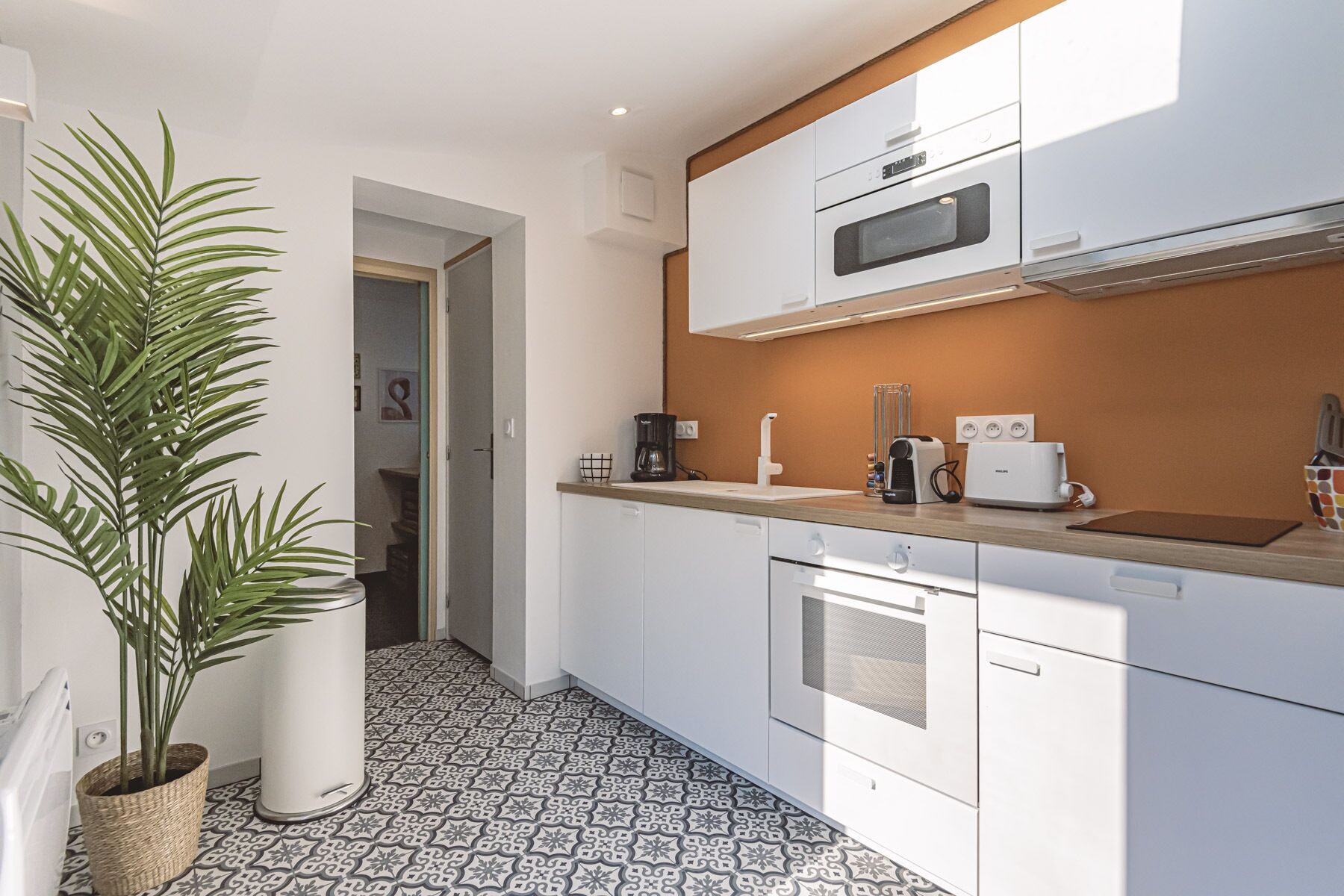 A modern kitchen with white cabinets, a microwave, a coffee maker, and a toaster. There is a potted plant on the left and a tiled floor.