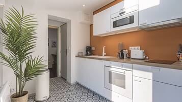 A modern kitchen with white cabinets, a microwave, a coffee maker, and a toaster. There is a potted plant on the left and a tiled floor.