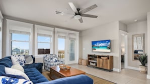 Een woonkamer met een blauwe bank, een houten salontafel, een plafondventilator, een televisie en uitzicht op het strand door grote ramen.